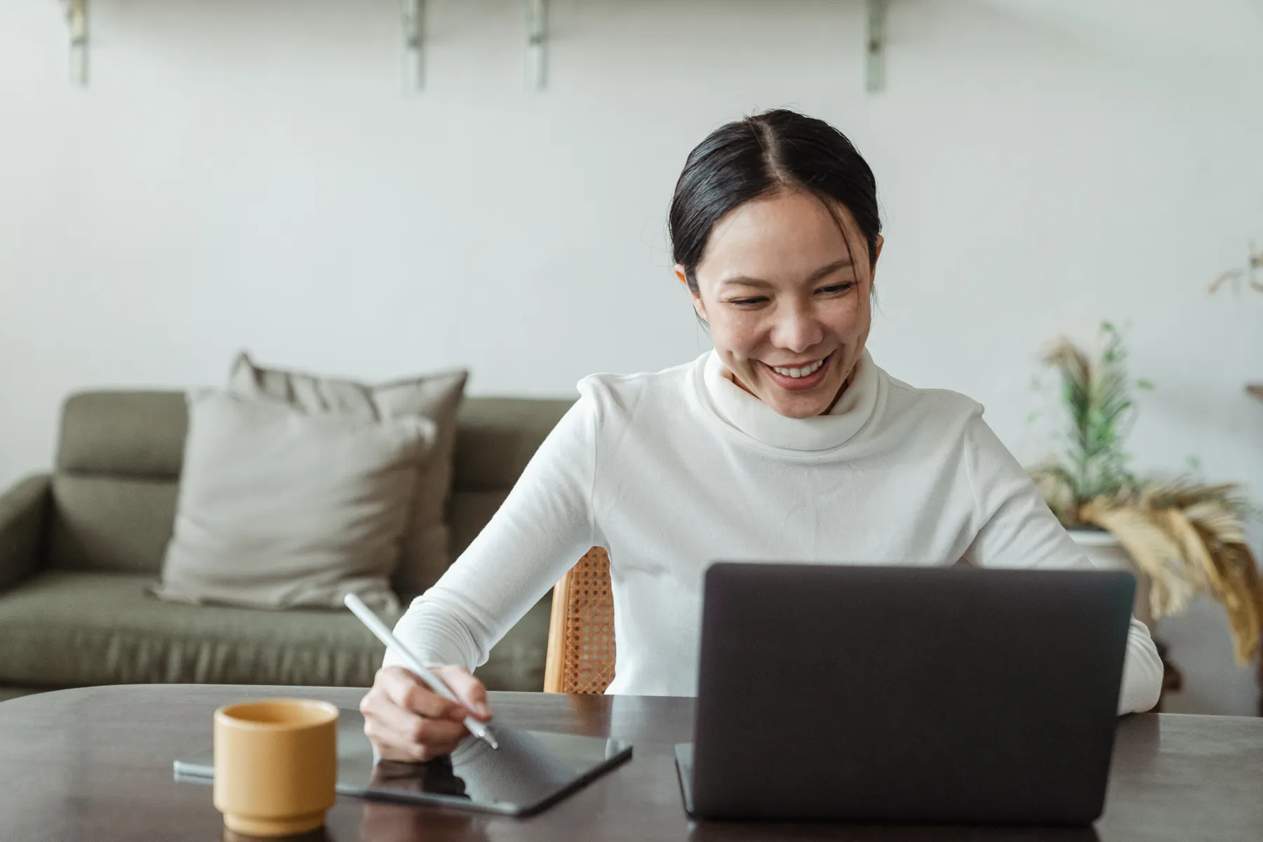 Mulher sorrindo ao olhar para um notebook enquanto faz anotações em um tablet em casa.